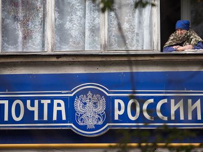 Пожилая женщина в окне своего балкона на фоне вывески "Почта России". Фото: Сафрон Голиков / Коммерсант